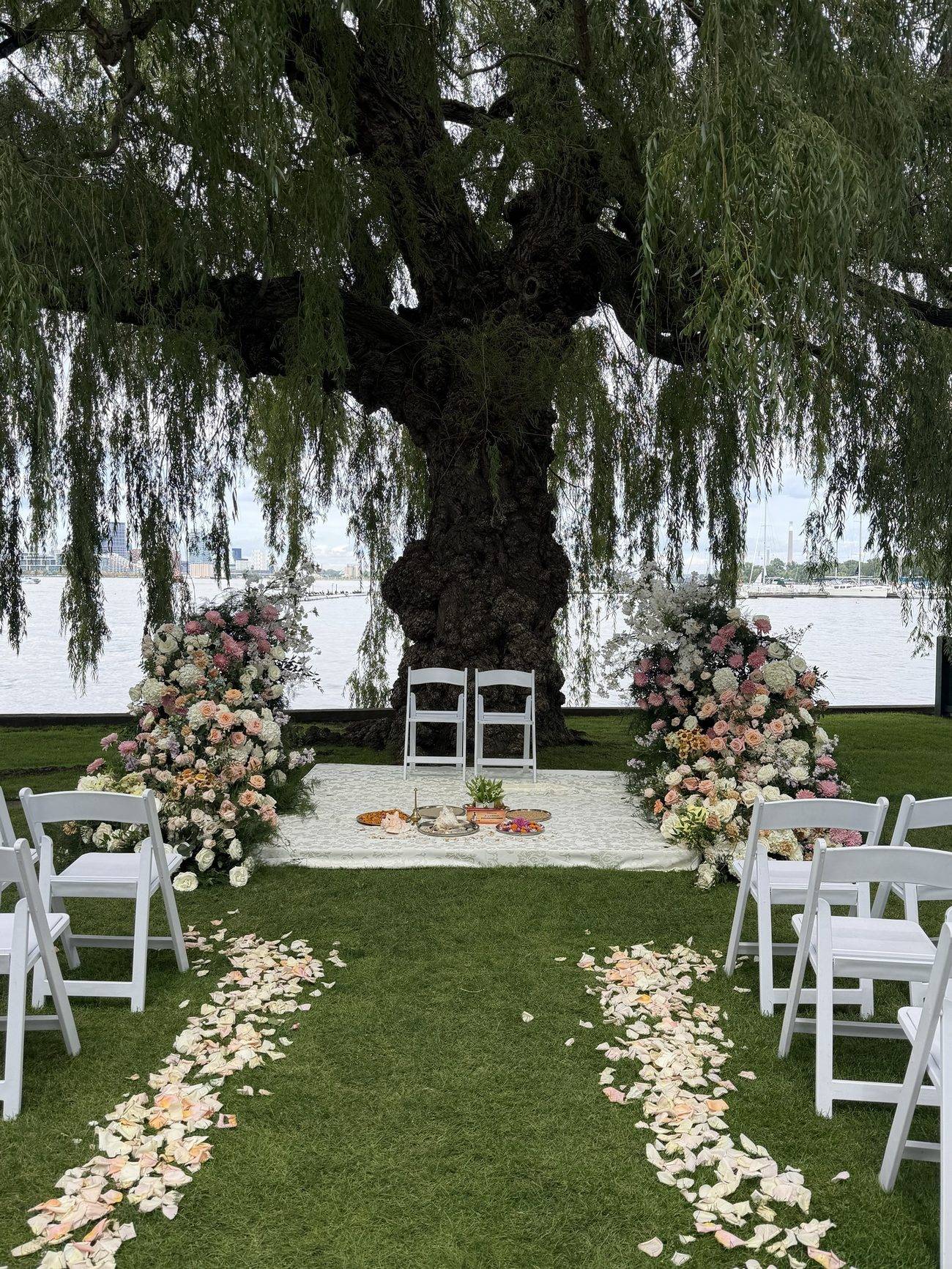 An outdoor waterside ceremony setup beneath a willow tree, with floral arrangements and a cultural ceremony table
