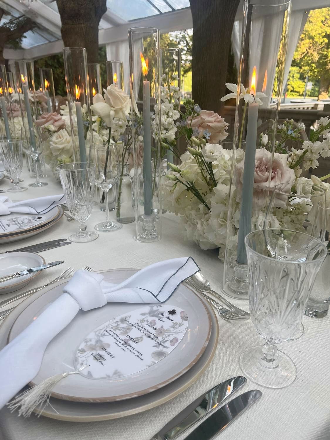 A long reception table set under a glass pavilion with tall taper candles and blush florals